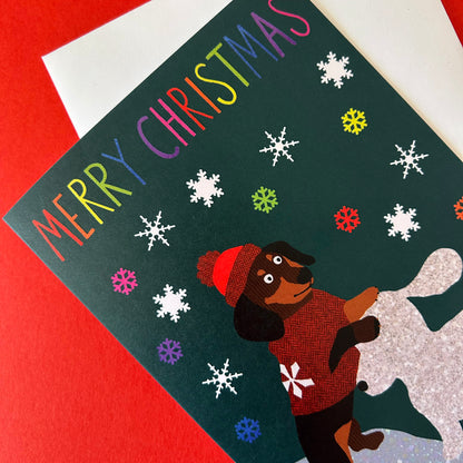 A Christmas greeting card featuring the text 'MERRY CHRISTMAS' in rainbow letters, decorated with white snowflakes on a dark green background. A dachshund wearing a red hat and red sweater is shown on a silver glittery surface.