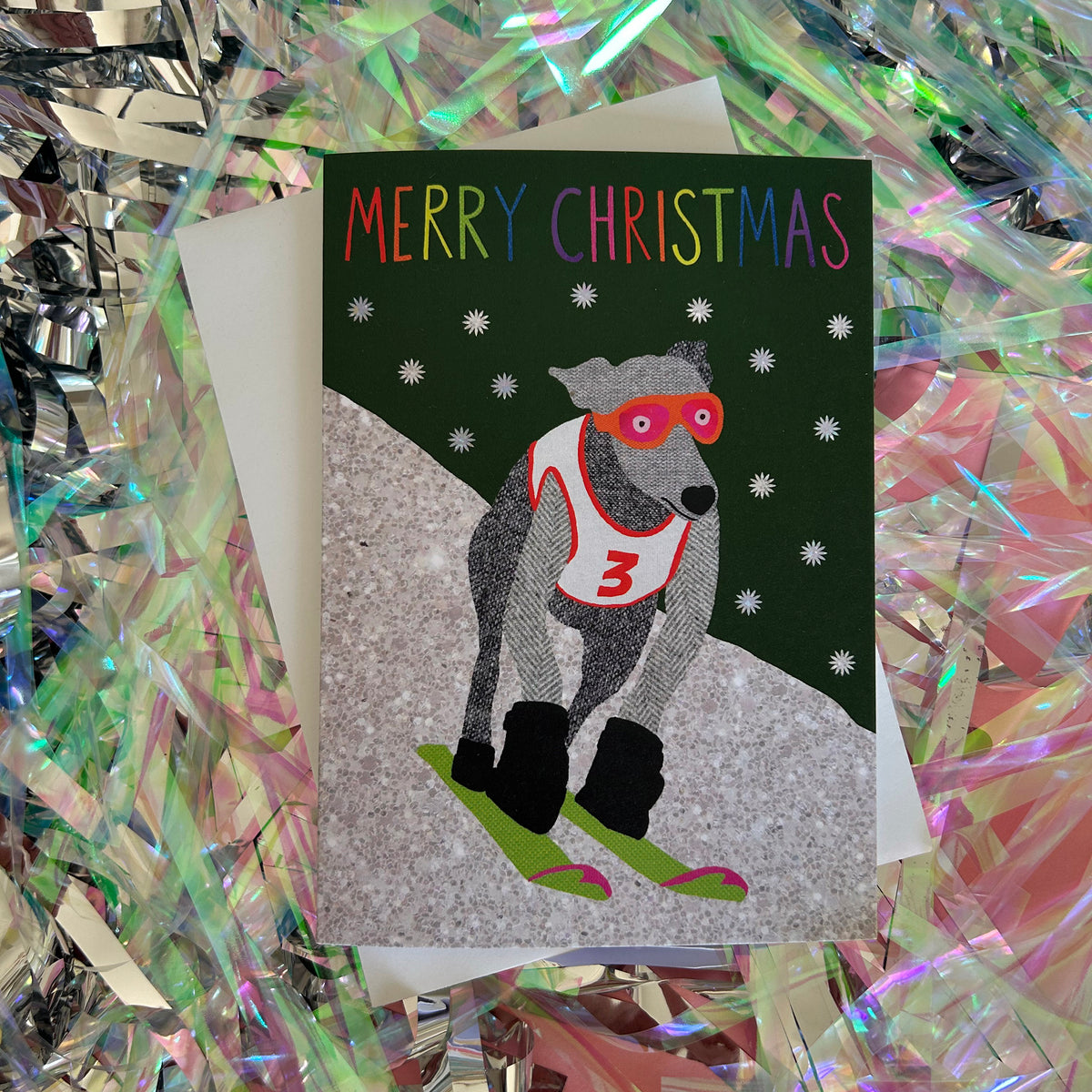 A Christmas greeting card featuring an  illustration of a grey dog wearing red goggles and a white racing number 3 jersey, skiing down a snowy hill. The card reads 'MERRY CHRISTMAS' in multicoloured letters against a dark green background with white snowflakes.