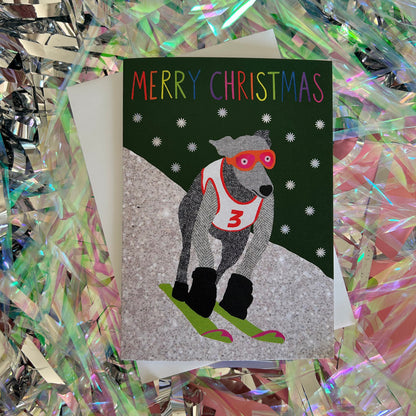 A Christmas greeting card featuring an  illustration of a grey dog wearing red goggles and a white racing number 3 jersey, skiing down a snowy hill. The card reads 'MERRY CHRISTMAS' in multicoloured letters against a dark green background with white snowflakes.