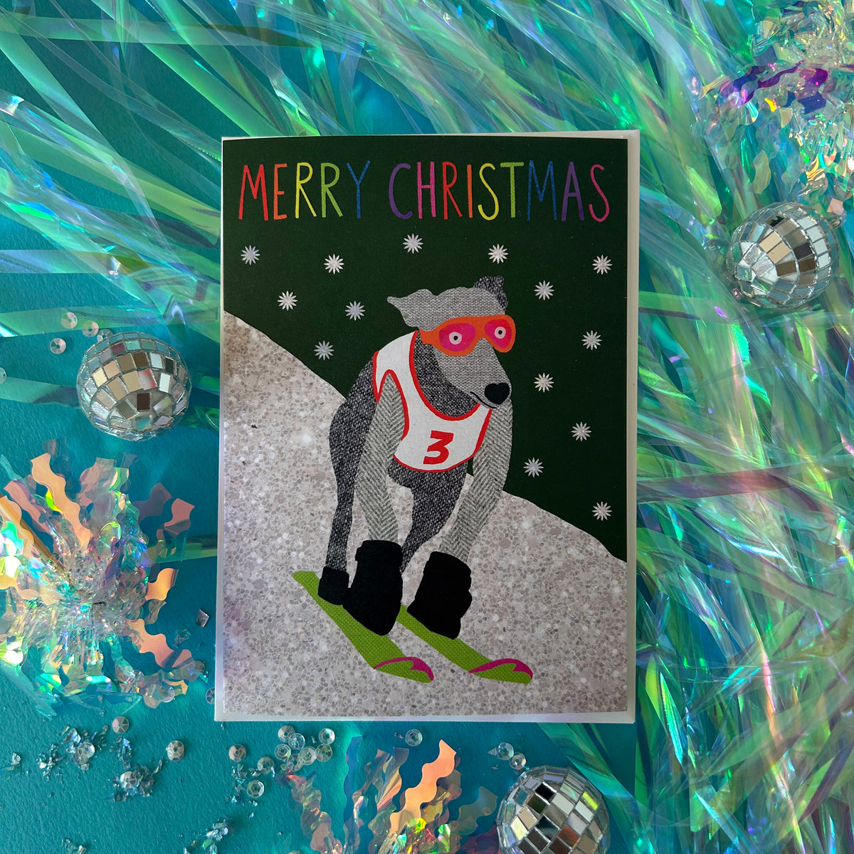A Christmas greeting card featuring a dog skier in a red outfit on green skis against a dark green background with snowflakes and the text 'MERRY CHRISTMAS' in rainbow letters. The card is displayed on a teal background with iridescent tinsel and small disco ball props.