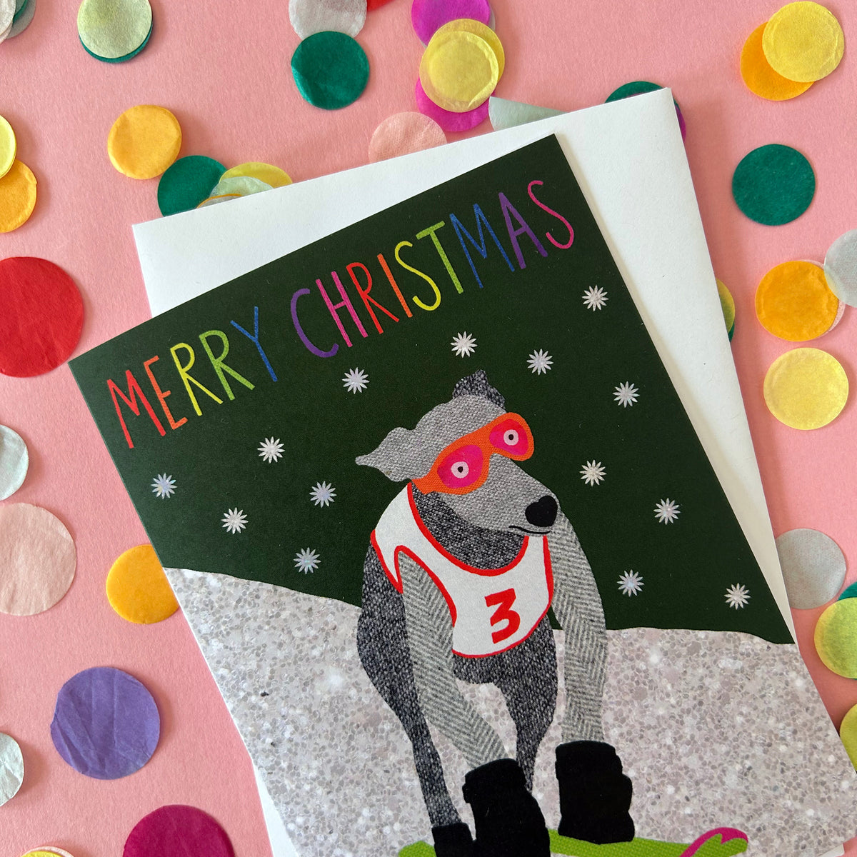 A festive Christmas greeting card featuring a greyhound dog wearing red goggles and a white jersey with the number 3, set against a dark green snowy background. The card is displayed on top of a white envelope, surrounded by colourful confetti on a pink background.