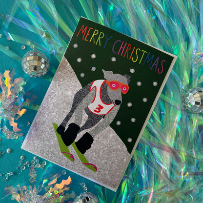 A festive Christmas greeting card featuring a greyhound wearing red goggles and a white jersey with the number 3, skiing down snowy hills. The card is styled on a teal background with holographic tinsel and mini disco balls.