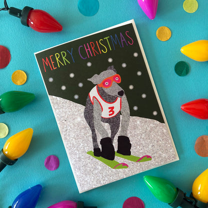 A Christmas greeting card featuring a dog wearing ski goggles and a racing bib with the number 3, set against a snowy background with the words 'MERRY CHRISTMAS' at the top. The card is styled in a flat-lay on a bright turquoise surface surrounded by colourful Christmas lights and confetti.