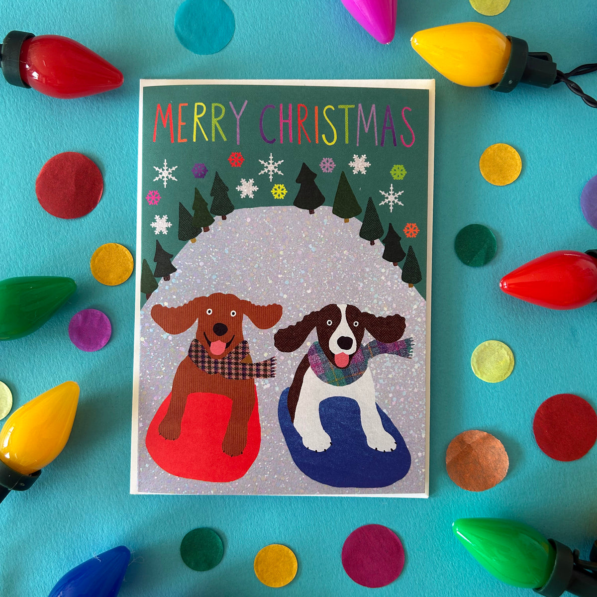 A Christmas greeting card featuring two dogs in a snowy scene with the text 'MERRY CHRISTMAS' at the top, styled on a bright turquoise background with colourful Christmas lights and confetti.