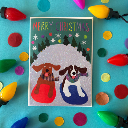 A Christmas greeting card featuring two dogs in a snowy scene with the text 'MERRY CHRISTMAS' at the top, styled on a bright turquoise background with colourful Christmas lights and confetti.