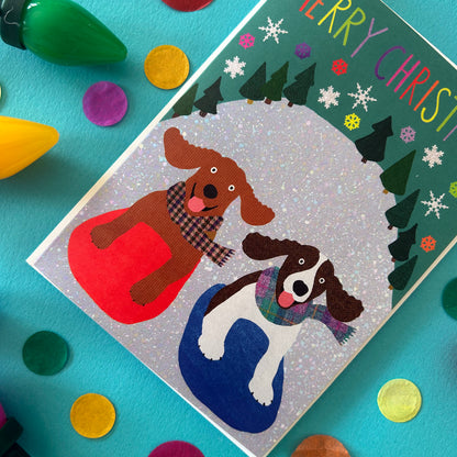 A Christmas card featuring two dogs sitting in red and blue sledges, decorated with glitter and colourful festive elements, shown on a teal background with colourful confetti and light bulbs.