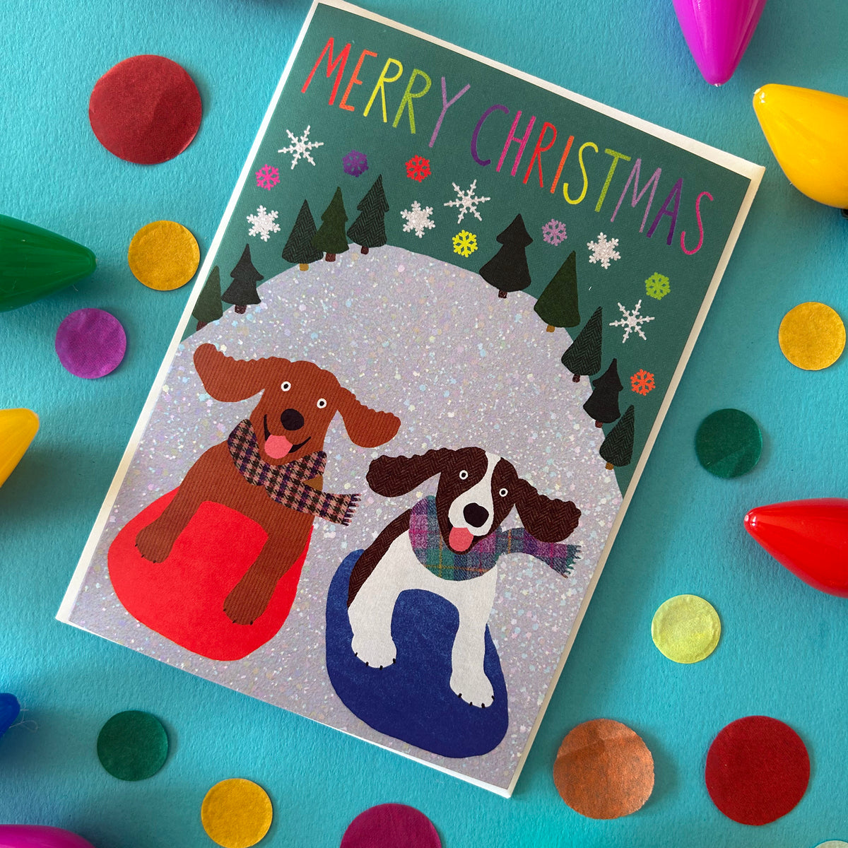 A hand drawn Christmas card featuring two dogs sitting in red and blue snow boots under a glittery white arch, surrounded by colourful felt circles and party shapes on a turquoise background.
