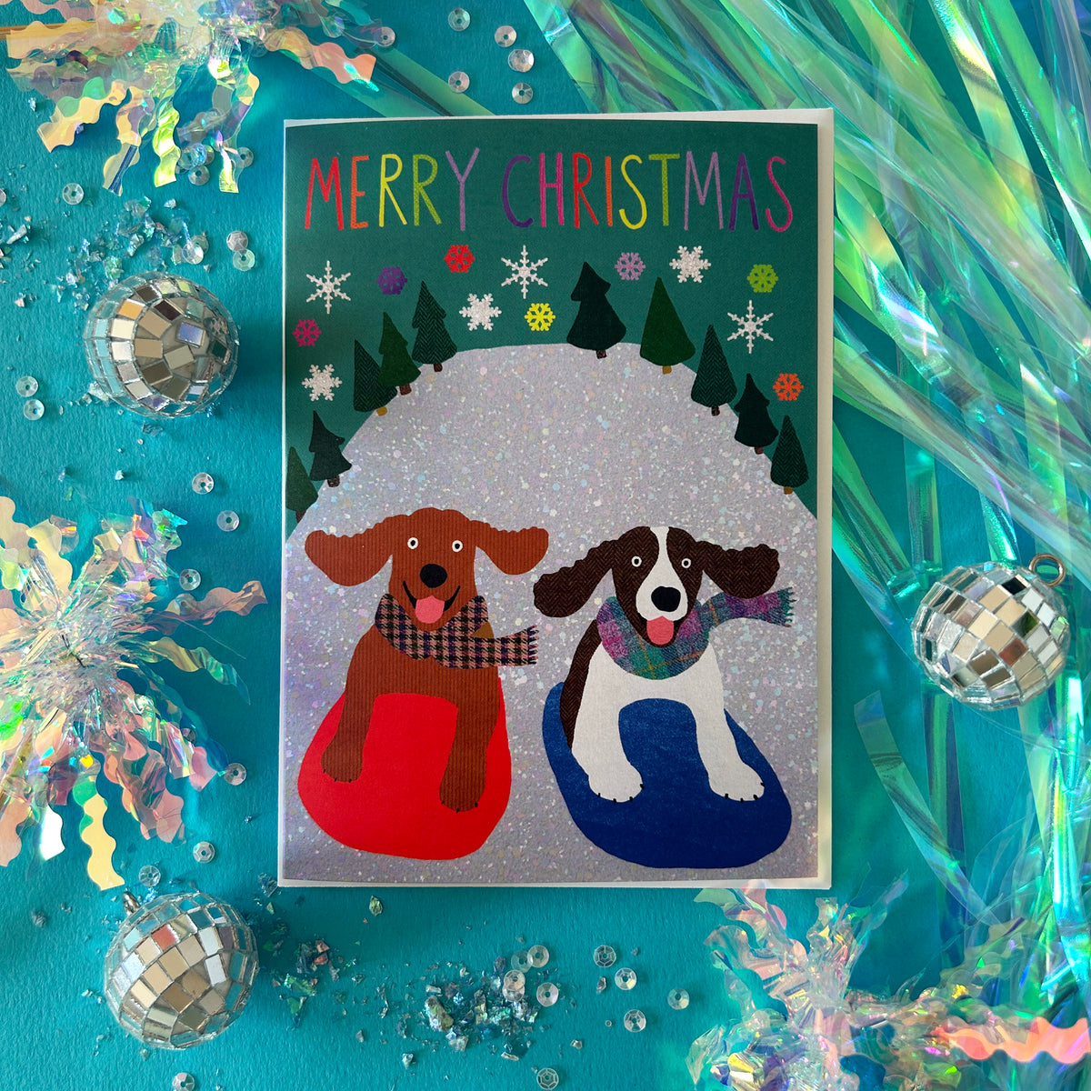 A bright Christmas greeting card featuring two dogs sitting in red and blue snow on a glittery background, surrounded by festive tinsel, disco balls, and sequins on a bright turquoise surface.