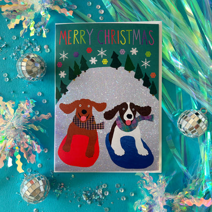 A bright Christmas greeting card featuring two dogs sitting in red and blue snow on a glittery background, surrounded by festive tinsel, disco balls, and sequins on a bright turquoise surface.