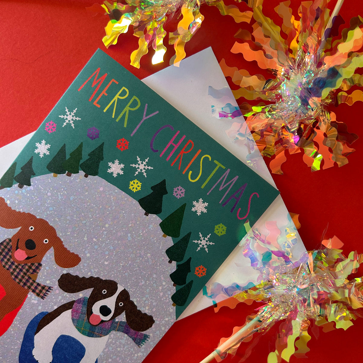 A Christmas greeting card featuring two dogs in winter scarves  sledging down a hill, with 'MERRY CHRISTMAS' in colourful lettering. The card is styled on a red background with festive tinsel decorations.