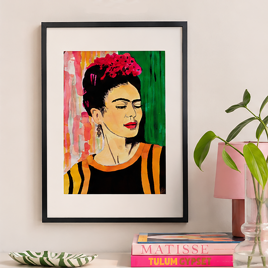 A framed art print featuring a colourful portrait of Frida Kahlo, styled on a wall above a stack of books and next to a pink lamp and green plant.