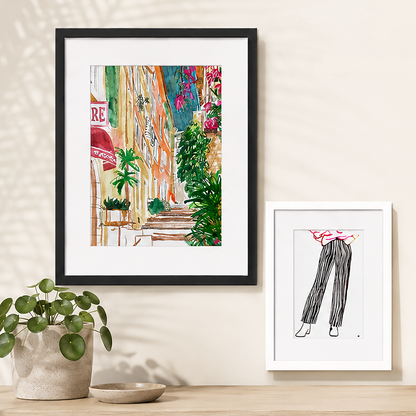 Two framed art prints displayed on a beige wall above a wooden surface. The larger left print has a black frame and white mat, showing a colourful Provence scene with green plants and warm-toned buildings.