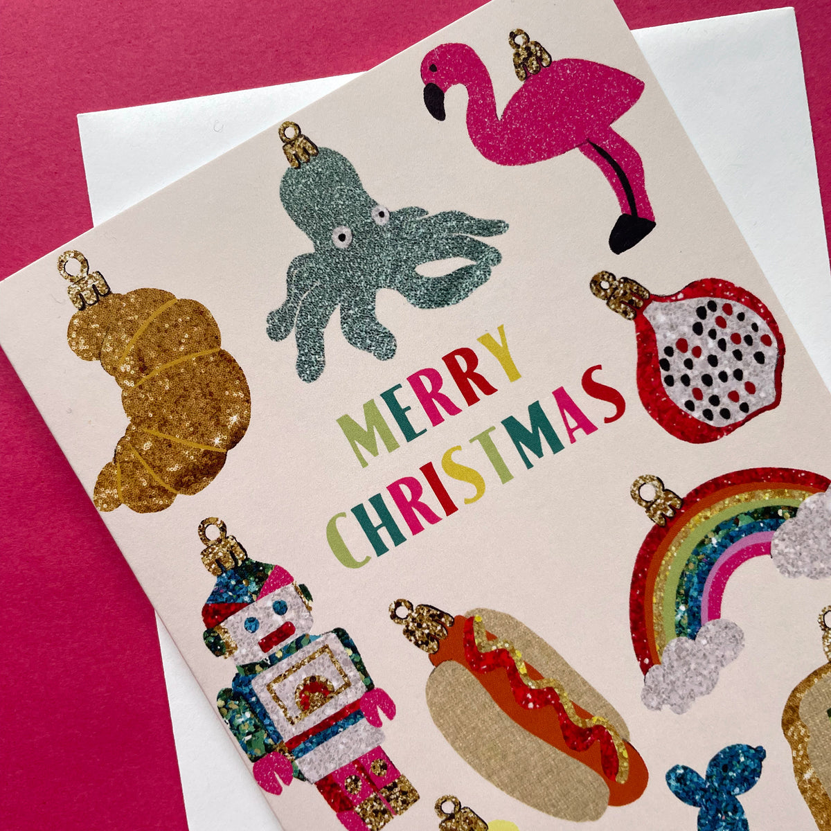 A Christmas greeting card featuring colourful glittery ornament illustrations and the words “MERRY CHRISTMAS” in multicoloured letters, placed on top of a white envelope against a pink background.