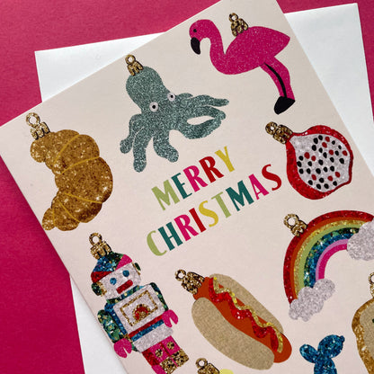 A Christmas greeting card featuring colourful glittery ornament illustrations and the words “MERRY CHRISTMAS” in multicoloured letters, placed on top of a white envelope against a pink background.