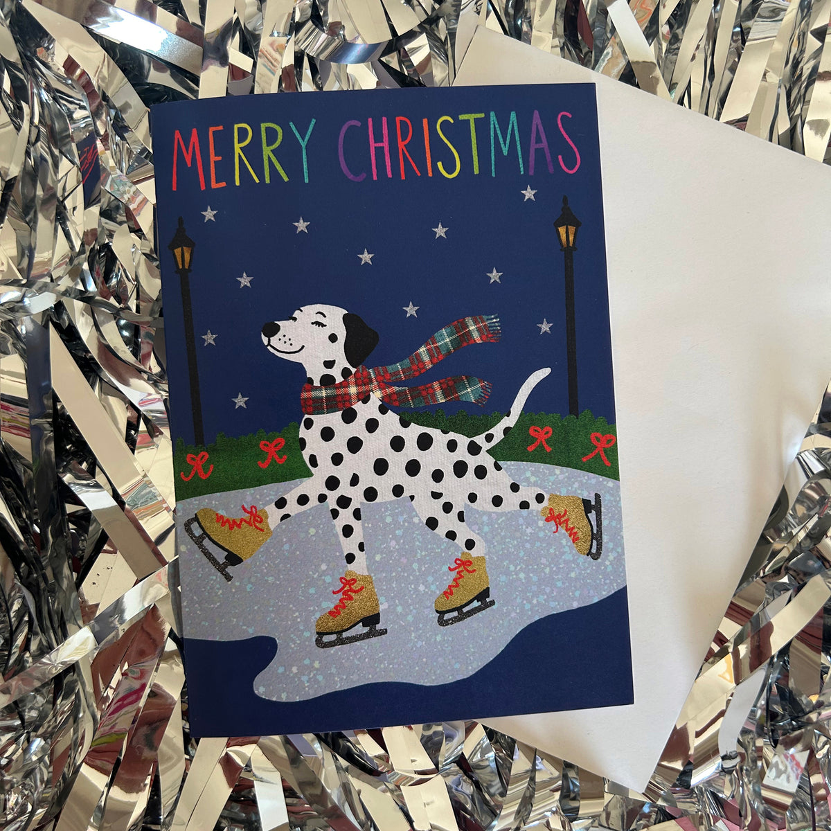 A Christmas greeting card featuring a Dalmatian ice skating in the snow under a dark blue sky with 'MERRY CHRISTMAS' written in colourful letters. The card is displayed on top of a white envelope, surrounded by shiny silver tinsel.