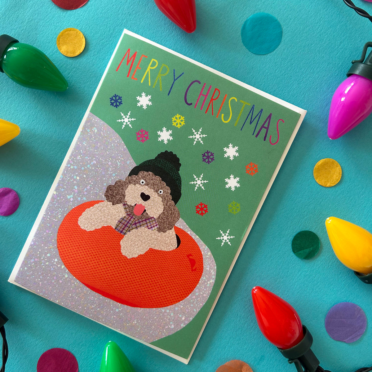 A Christmas greeting card featuring a fluffy cockapoo dog sitting in an orange inner tube on a green background with snowflakes and the text 'MERRY CHRISTMAS'. The card is styled in a flat-lay on a bright blue surface surrounded by colourful Christmas lights and confetti.
