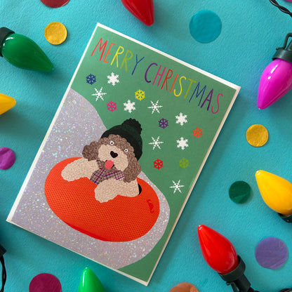 A Christmas greeting card featuring a fluffy cockapoo dog sitting in an orange inner tube on a green background with snowflakes and the text 'MERRY CHRISTMAS'. The card is styled in a flat-lay on a bright blue surface surrounded by colourful Christmas lights and confetti.