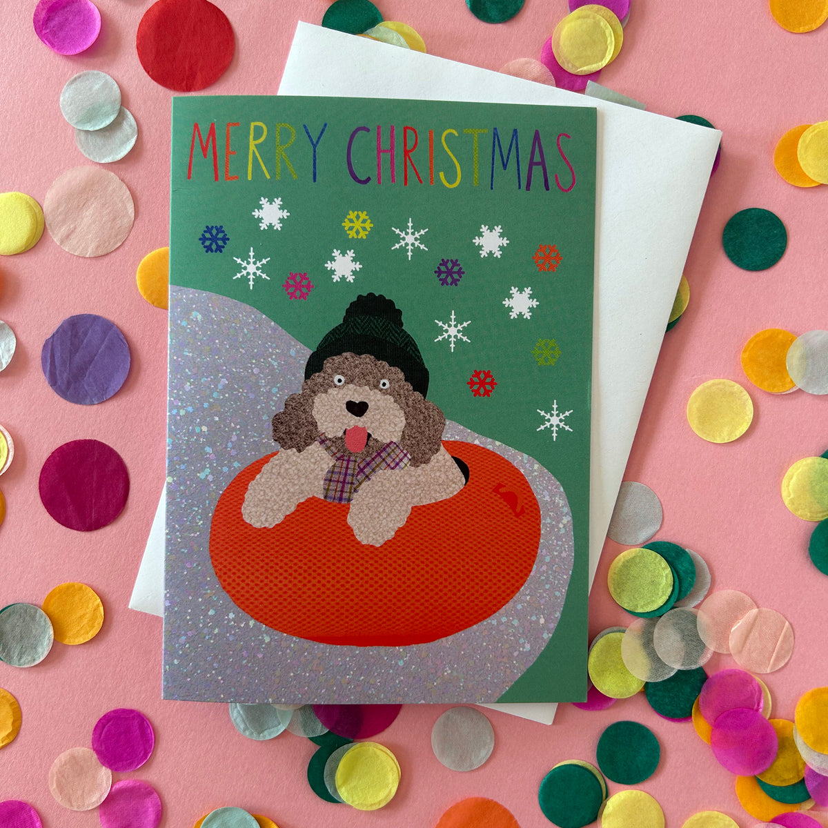 A Christmas greeting card featuring a dog in a red inner tube on a green background with snowflakes and the text 'MERRY CHRISTMAS'. The card is layered over a white envelope and surrounded by colourful confetti on a pink surface.