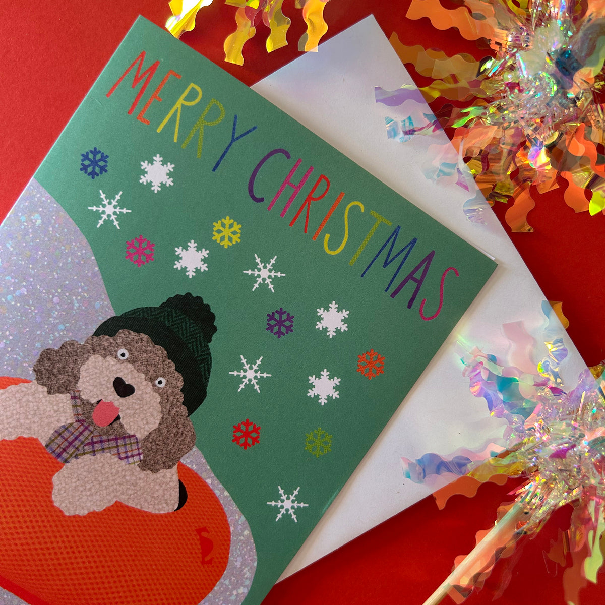 A Christmas greeting card featuring a green background, colourful 'MERRY CHRISTMAS' text, snowflakes, and a dog illustration wearing a hat and plaid scarf, styled on a red surface with a white envelope and iridescent tinsel.