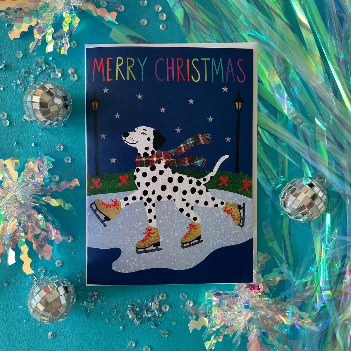 A Christmas greeting card featuring a Dalmatian dog ice skating in a snowy park under a dark blue night sky, surrounded by festive iridescent tinsel, disco balls, and silver ornaments on a turquoise background.