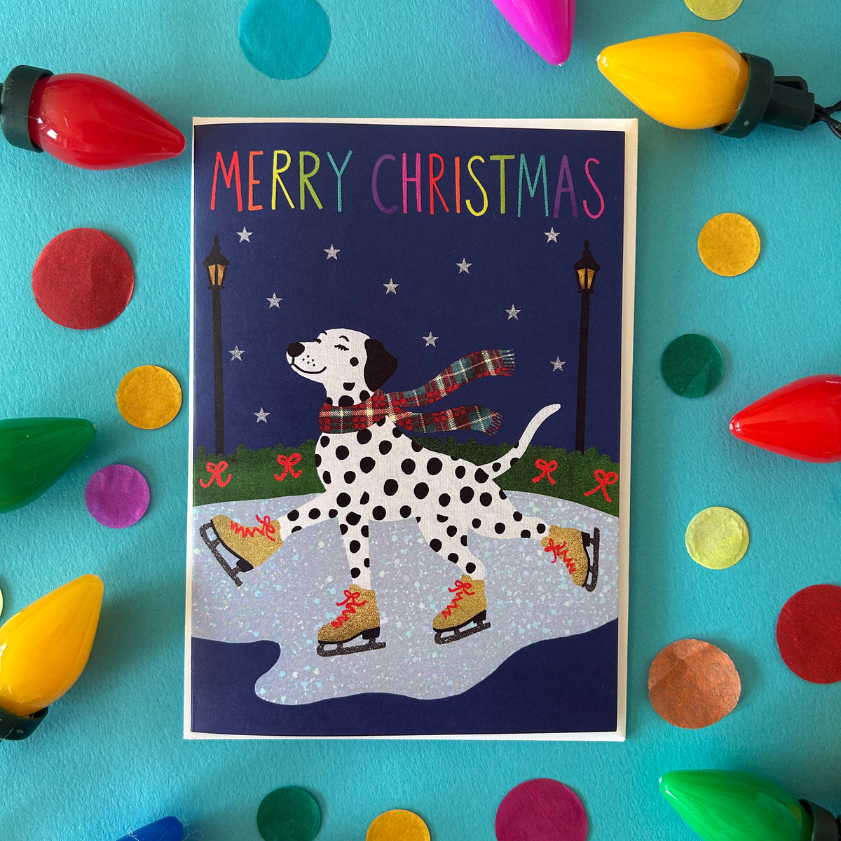 A Christmas greeting card featuring a Dalmatian dog ice skating in a snowy park at night, surrounded by colourful paper confetti and decorative Christmas bulb lights on a bright turquoise background.