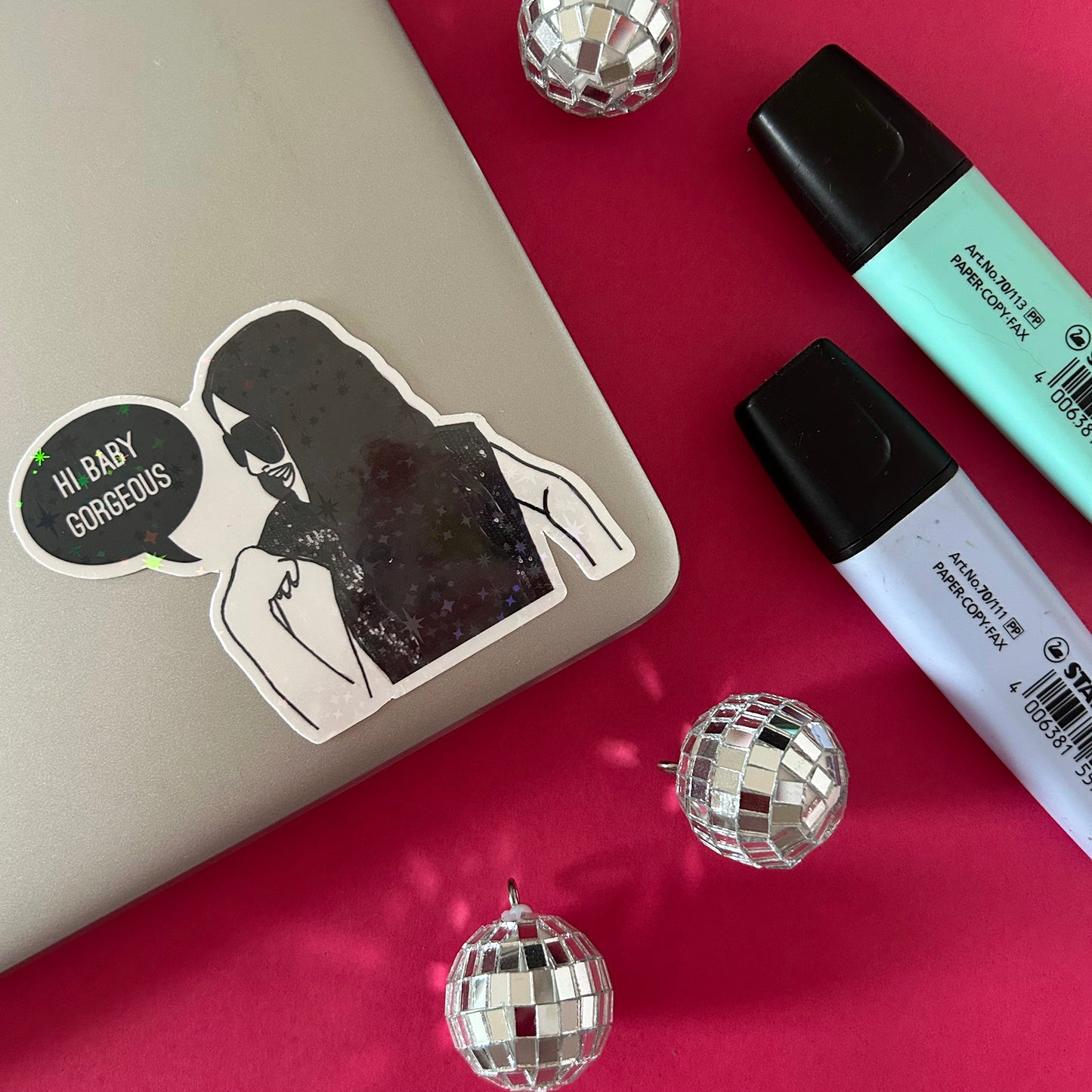 Sticker with a design of RHOSLC's Lisa saying 'Hi BabyGorgeous', on a laptop with markers, and disco balls on a red surface.