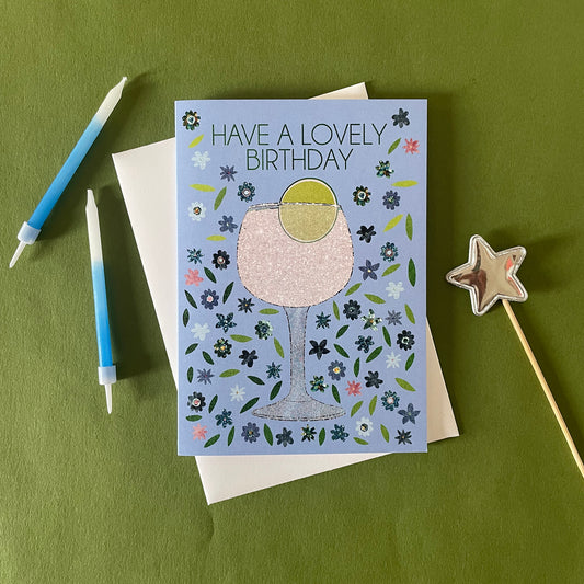 Image of a pastel blue birthday card featuring an illustration of a gin and tonic surrounded  by flowers and says  Have a Lovely Birthday