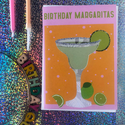 Image shows a yellow and pink coloured  birthday card with the greeting 'Birthday Margaritas' and a lime green cocktail with limes and a glitter salt rim.