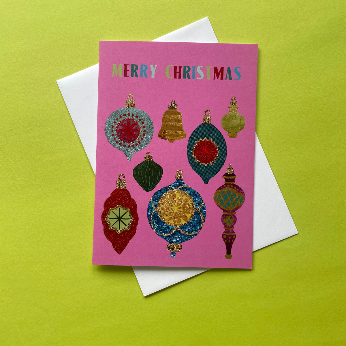 A pink Christmas greeting card with colourful ornament illustrations and the words 'MERRY CHRISTMAS' at the top, placed on top of a white envelope against a bright yellow-green background.