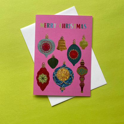 A pink Christmas greeting card with colourful ornament illustrations and the words 'MERRY CHRISTMAS' at the top, placed on top of a white envelope against a bright yellow-green background.