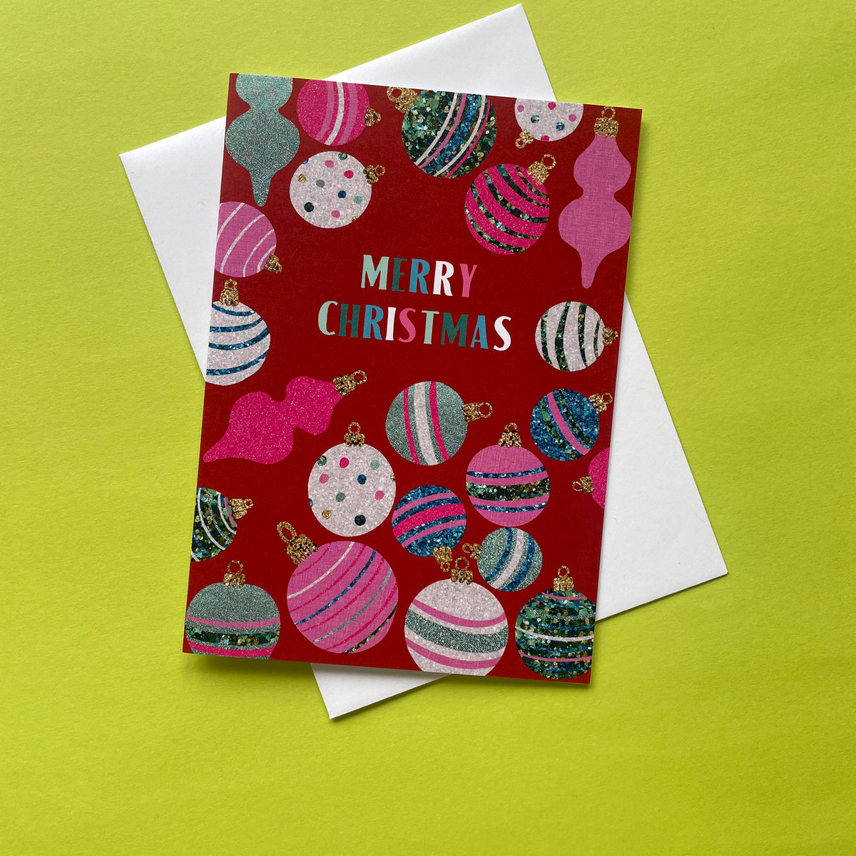 A red Christmas greeting card with a festive pattern of colourful striped and glittery ornaments, featuring the text 'MERRY CHRISTMAS' in white and pink. The card is displayed on top of a white envelope against a bright lime green background.