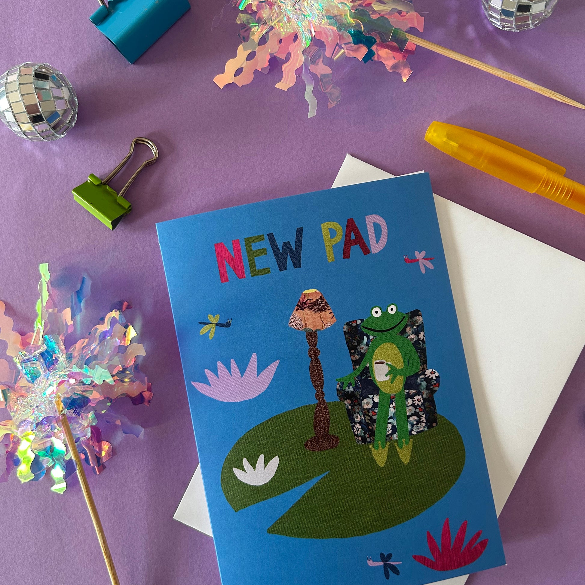 New home card with a frog illustration on a purple surface with party supplies