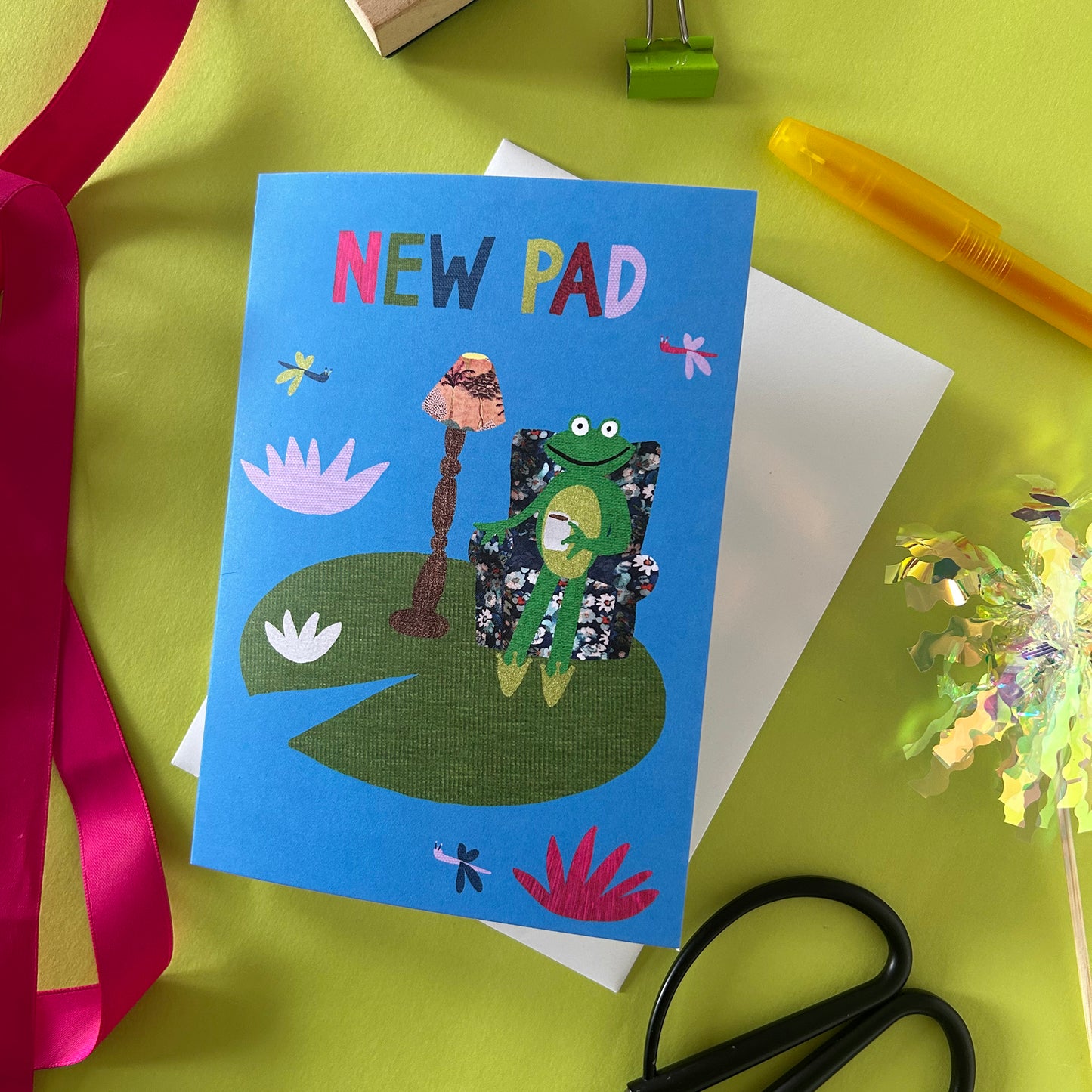Card with a frog design and 'NEW PAD' text on a green surface with stationery items.