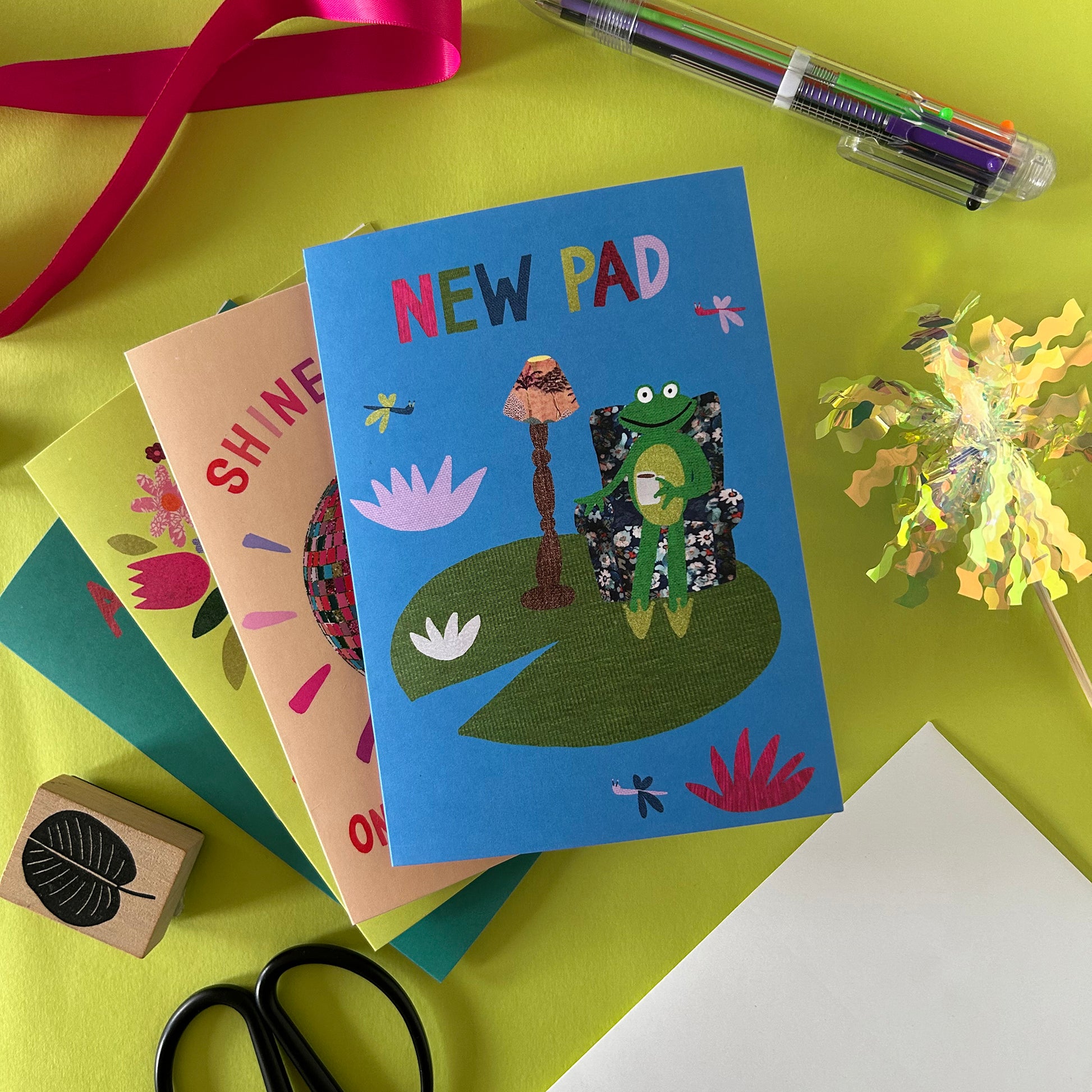 Colourful New Pad Card with a frog design on a green surface with stationery items.