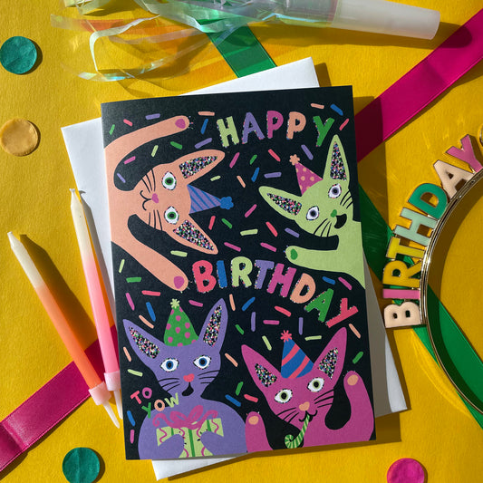 Image shows a quirky black greetings card with neon, illustrated cats in party hats and the words happy birthday.