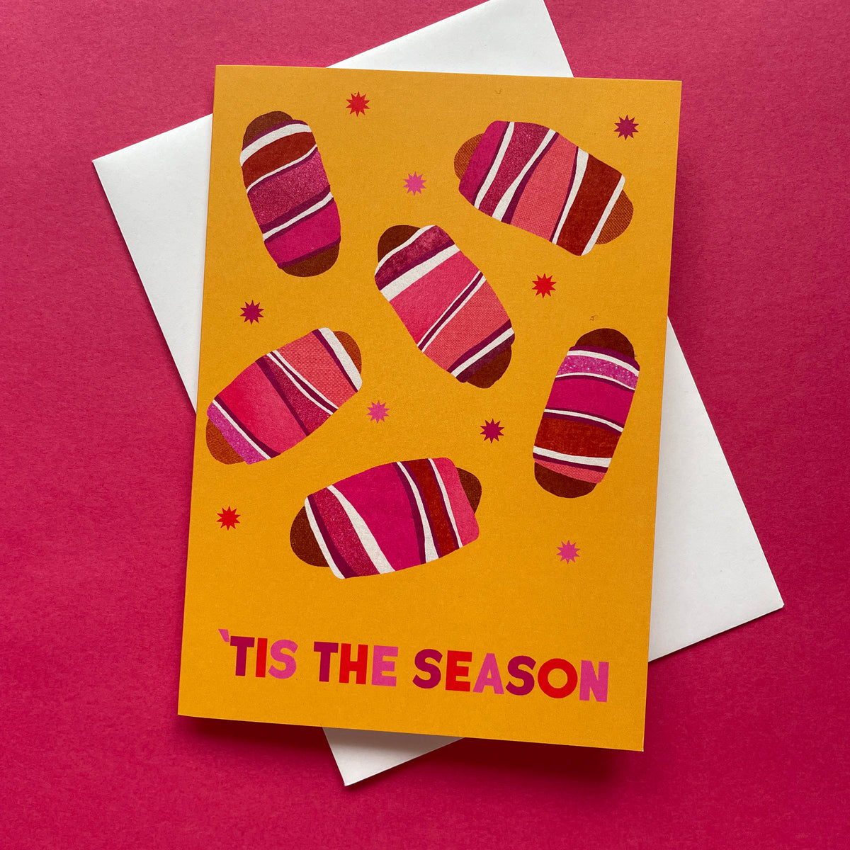 Greeting card with pigs in blankets design and 'Tis the Season text on a pink background