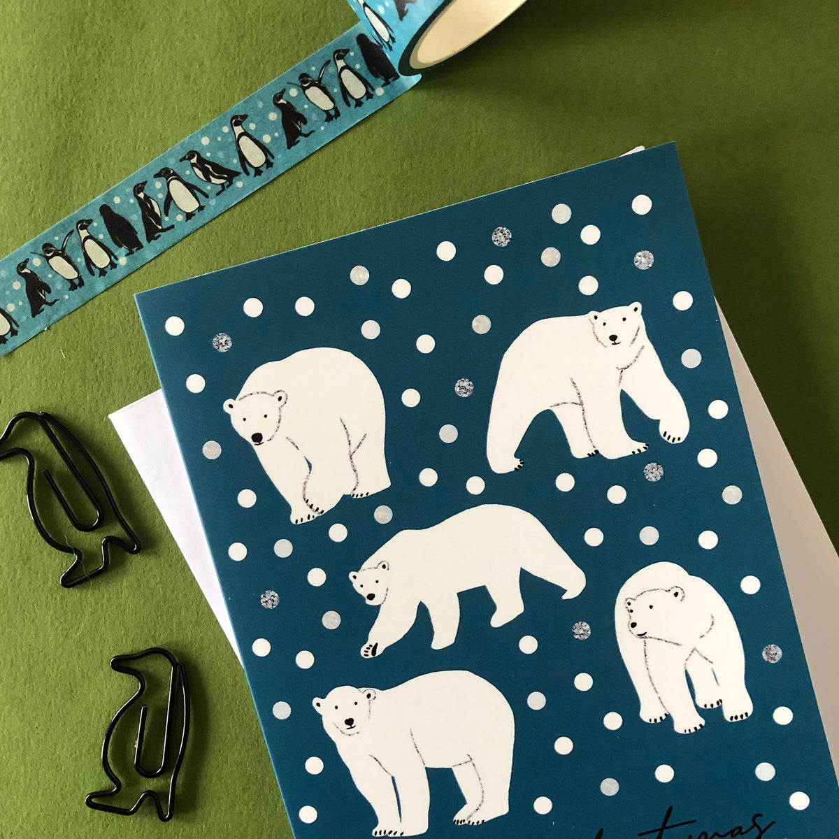 Arctic Animals greetings card featuring white polar bears on a dark teal-blue background with white snowballs, styled on a textured olive green surface with a penguin-patterned washi tape roll and black penguin-shaped clips.