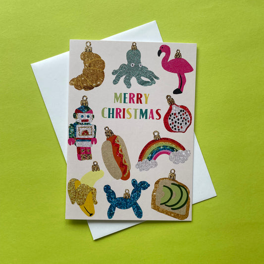 A Christmas ornament card featuring colourful novelty baubles placed on top of a white envelope against a bright yellow-green background.