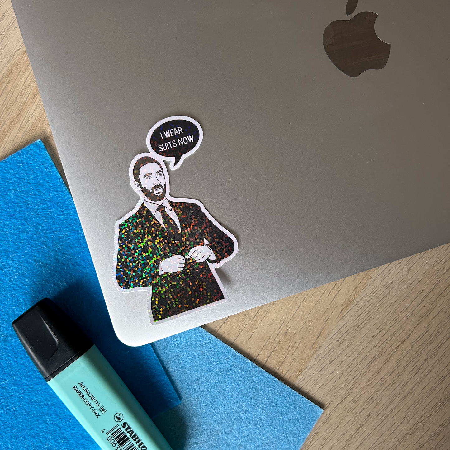 Laptop with a sticker of a man in a suit and a speech bubble saying 'I wear suits now' on a wooden surface.
