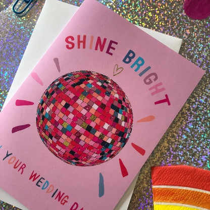 Image shows a pink wedding card featuring a glittery rainbow disco ball illustration and the words 'Shine Bright on your wedding day'.