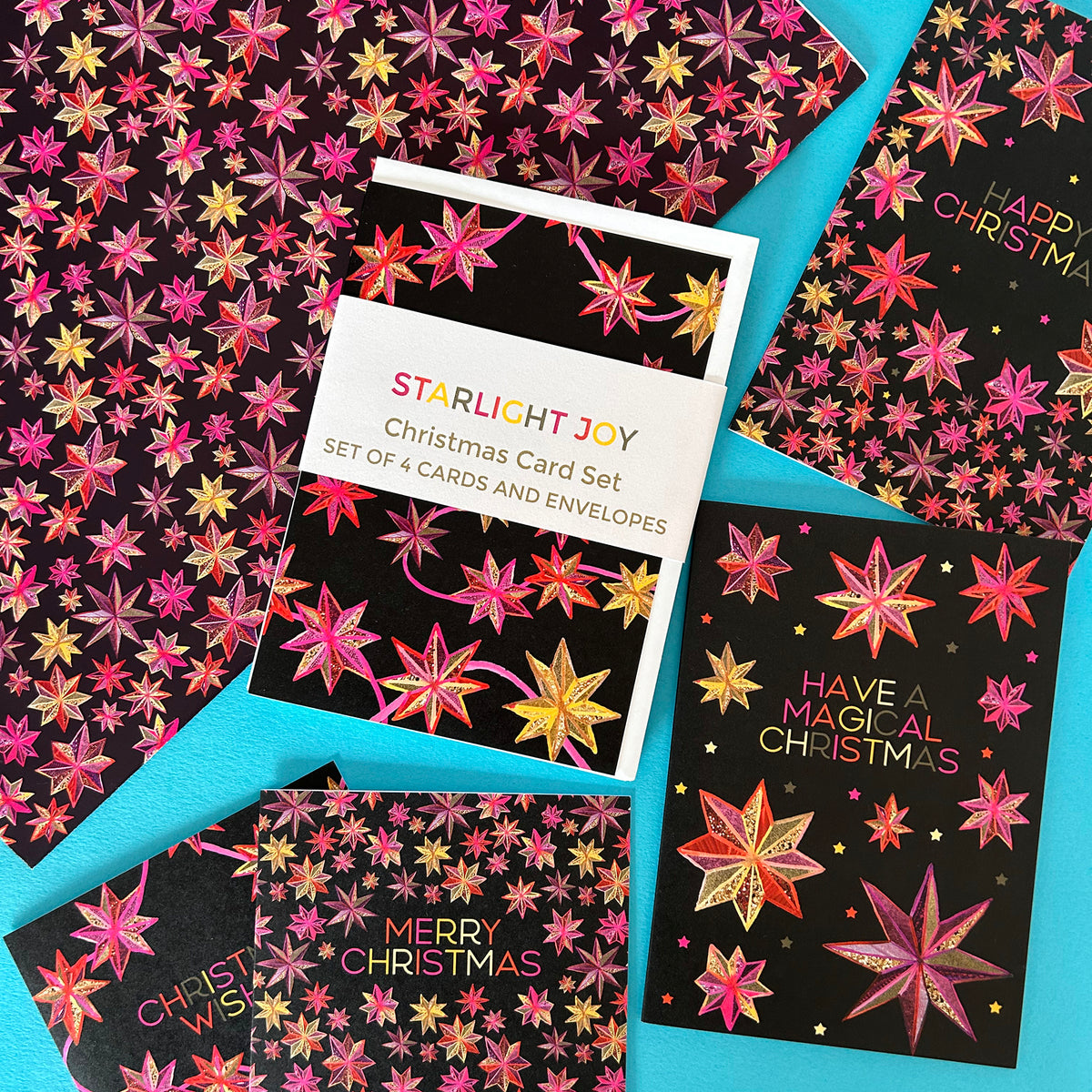 A flat-lay of a 'Starlight Joy' Christmas card set featuring multiple black cards and envelopes decorated with colourful pink, gold, and red star patterns, arranged on a bright turquoise background.