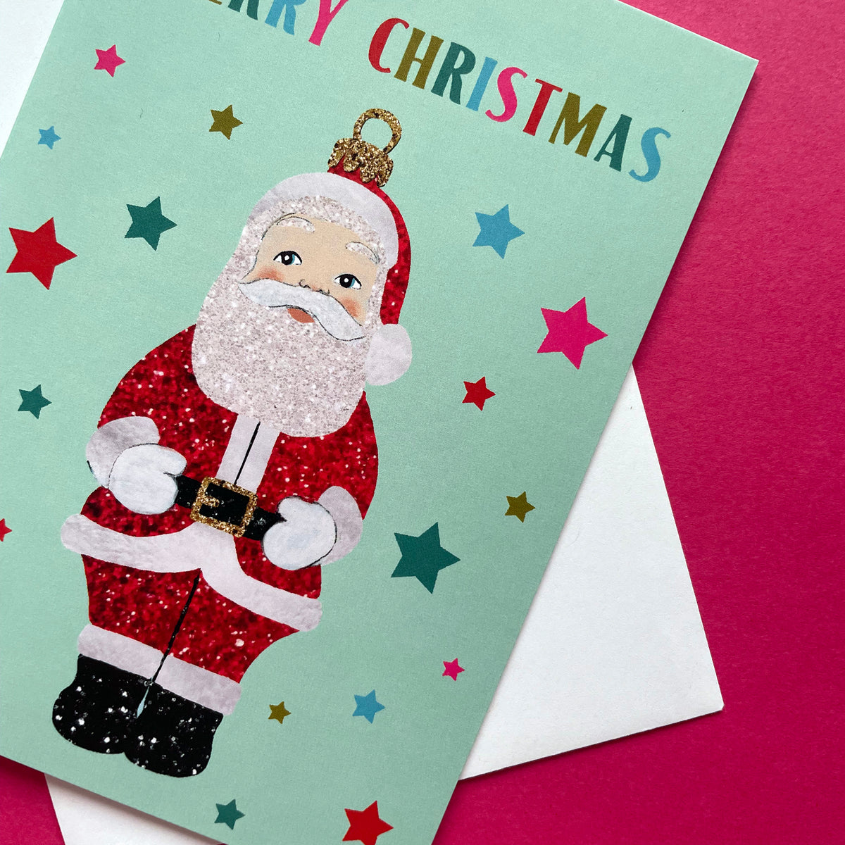 A festive Christmas greeting card featuring a glittery Santa Claus illustration on a mint green background with colourful stars and the text 'MERRY CHRISTMAS' at the top. The card is layered over a white envelope on a bright pink surface.