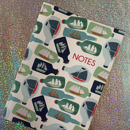 Image of a ships in bottles print A5 size notebook in a cream colour with blue and green bottles. 