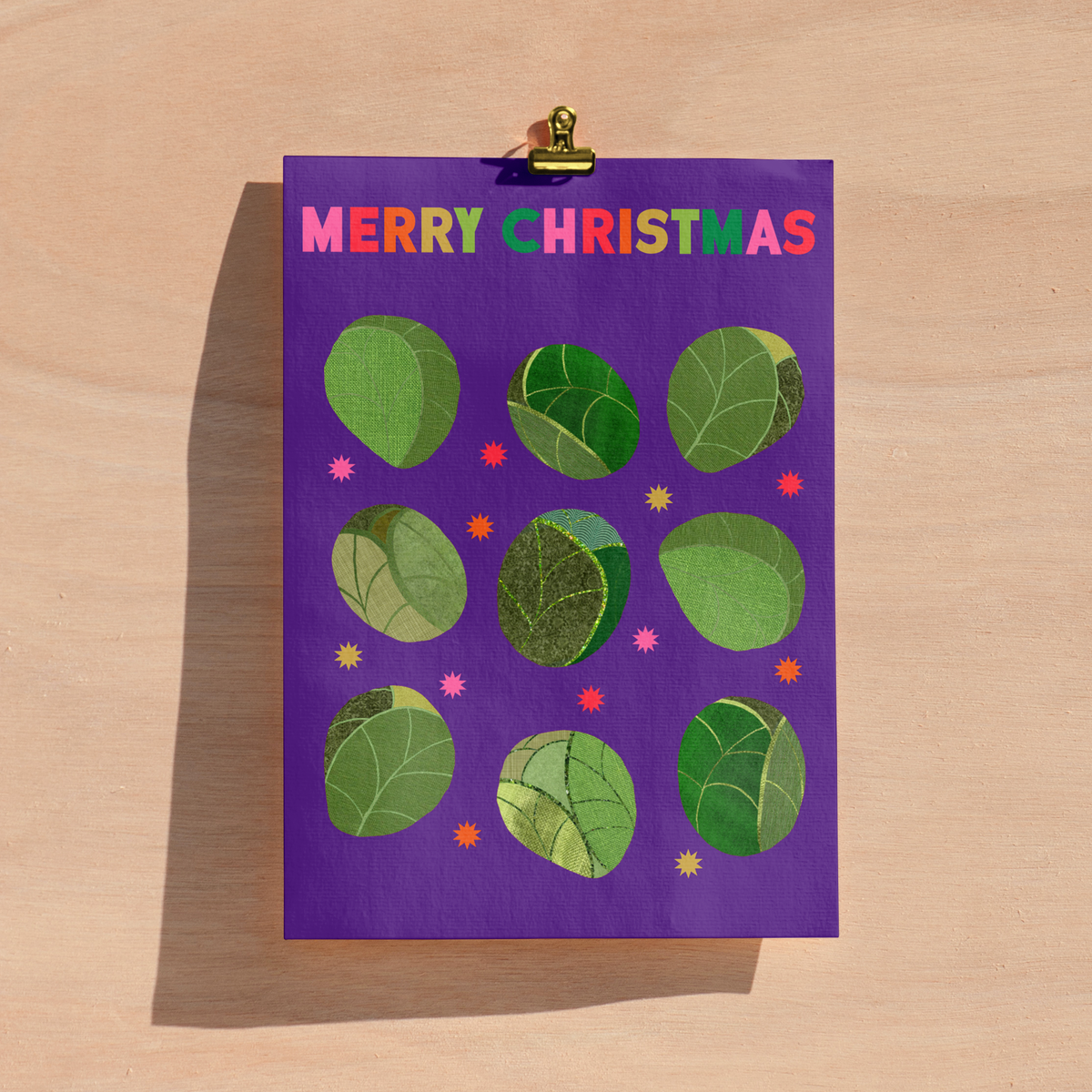 A purple Christmas greeting card with the text 'MERRY CHRISTMAS' in multicoloured letters, decorated with green sprout illustrations, shown clipped to a light wood background.