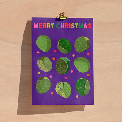 A purple Christmas greeting card with the text 'MERRY CHRISTMAS' in multicoloured letters, decorated with green sprout illustrations, shown clipped to a light wood background.
