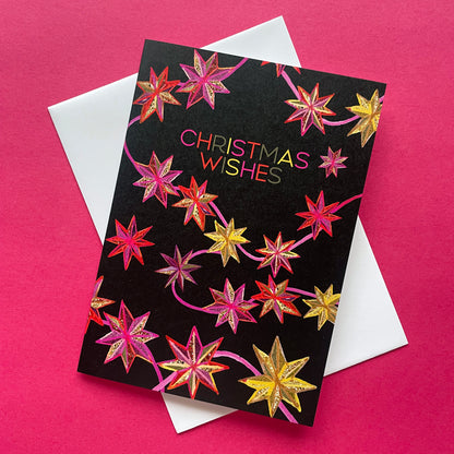 A black Christmas greeting card with  gold and pink starburst designs and the words 'CHRISTMAS WISHES' in multicolour lettering, shown with a white envelope on a bright pink background.