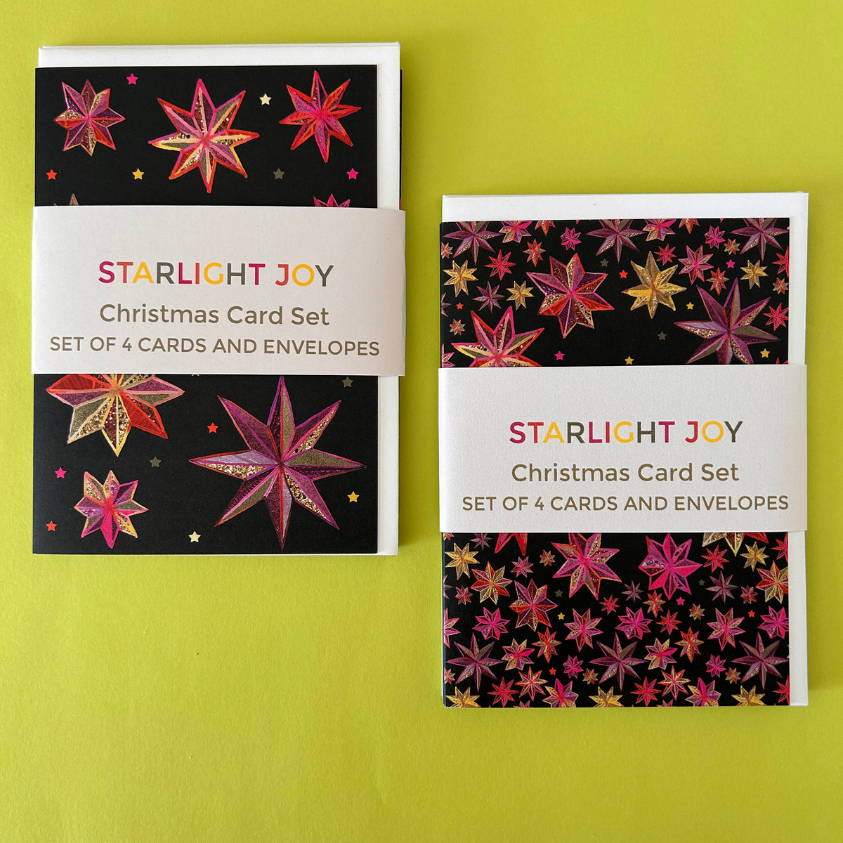 Two copies of the Starlight Joy Christmas Card Set displayed side-by-side on a bright yellow-green background. The set features black cards decorated with colourful starburst patterns in pink, red, and gold, complete with matching envelopes.