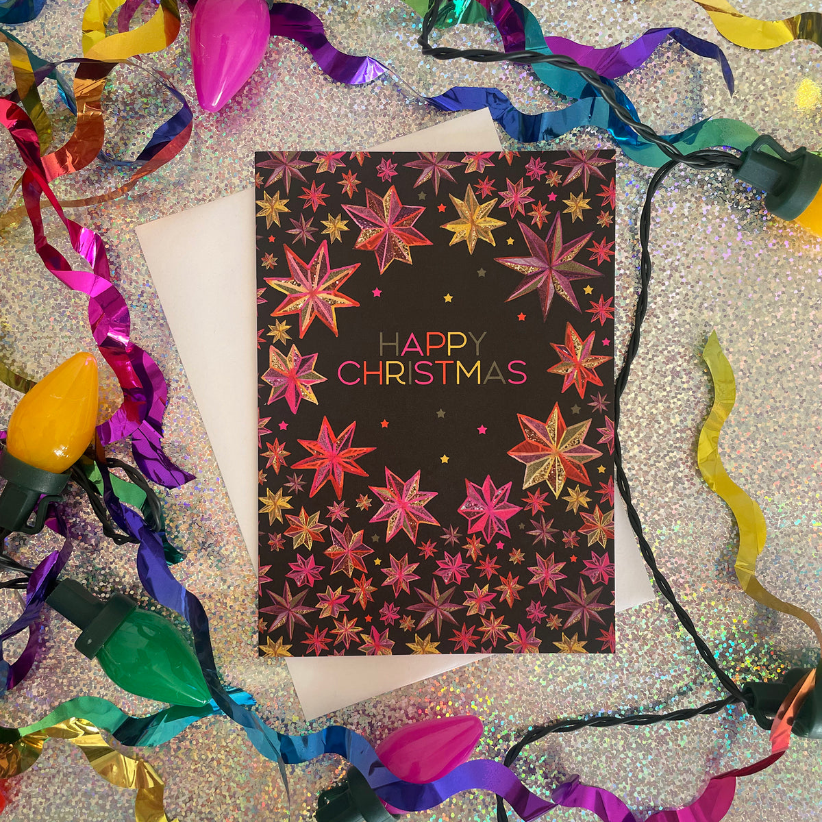 A black Christmas greeting card with a colourful star patterns, surrounded by multicoloured Christmas lights, ribbons, and a white envelope on a glittery silver surface.
