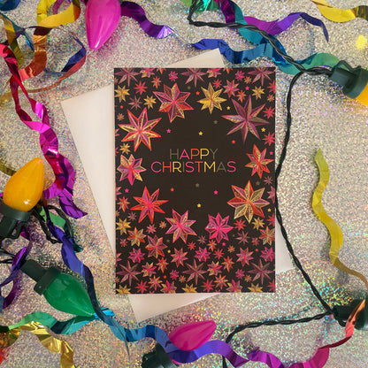 A black Christmas greeting card with a colourful star patterns, surrounded by multicoloured Christmas lights, ribbons, and a white envelope on a glittery silver surface.