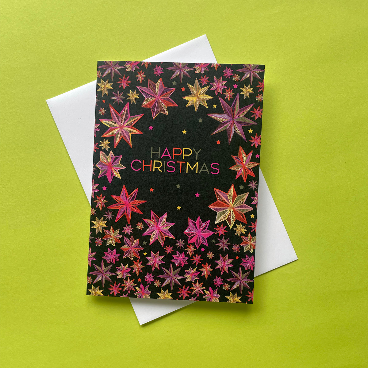 A bright, folk style Christmas greeting card with a black background and colourful glittery star pattern, featuring the words 'HAPPY CHRISTMAS' in pink and gold. The card is layered over a white envelope on a bright lime green background.